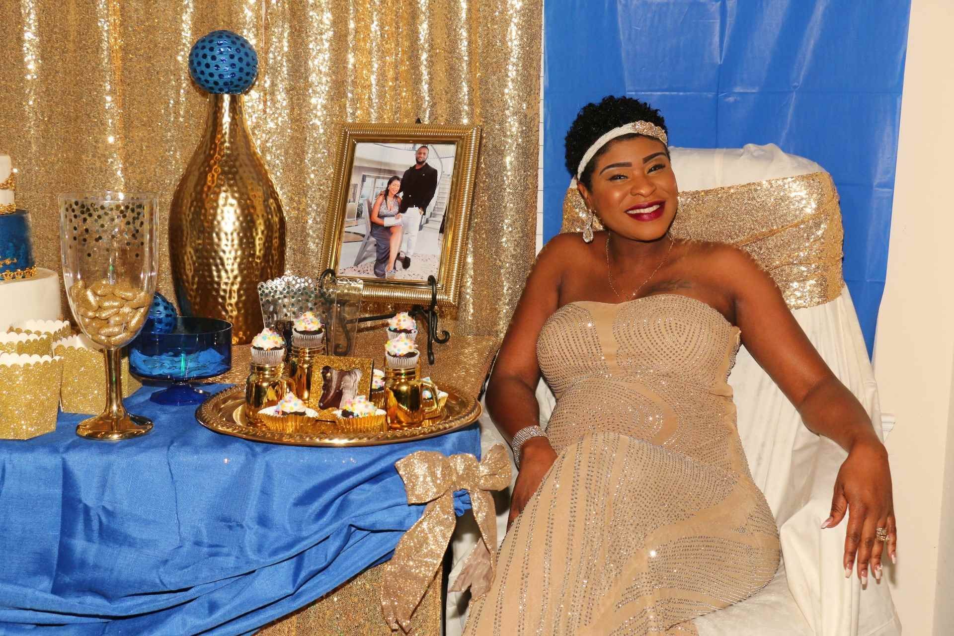 Woman in a gold gown smiles on a decorated chair beside a blue-and-gold table at a formal celebration.