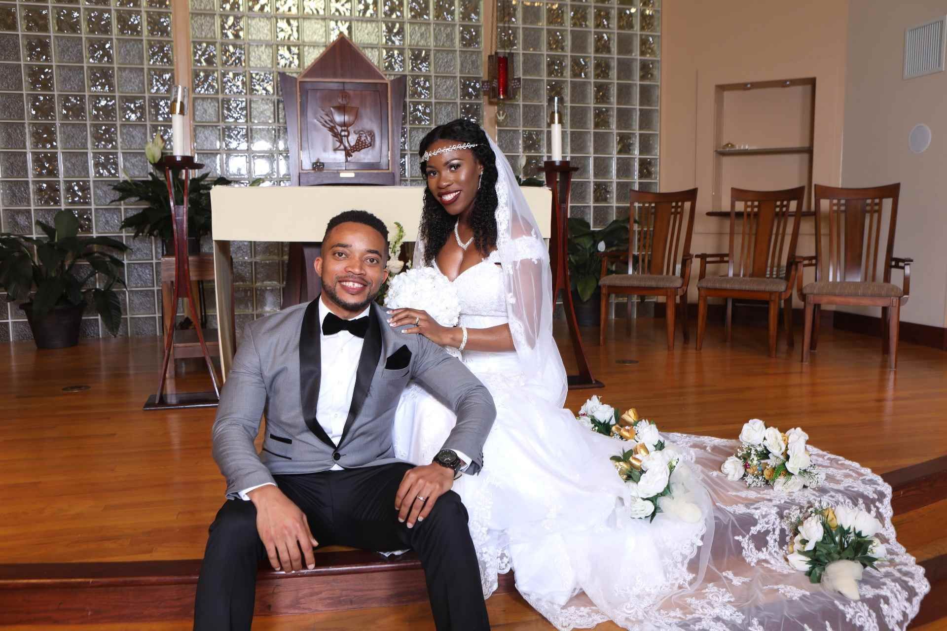 Bride and groom seated on church steps, smiling in wedding attire with white floral bouquet