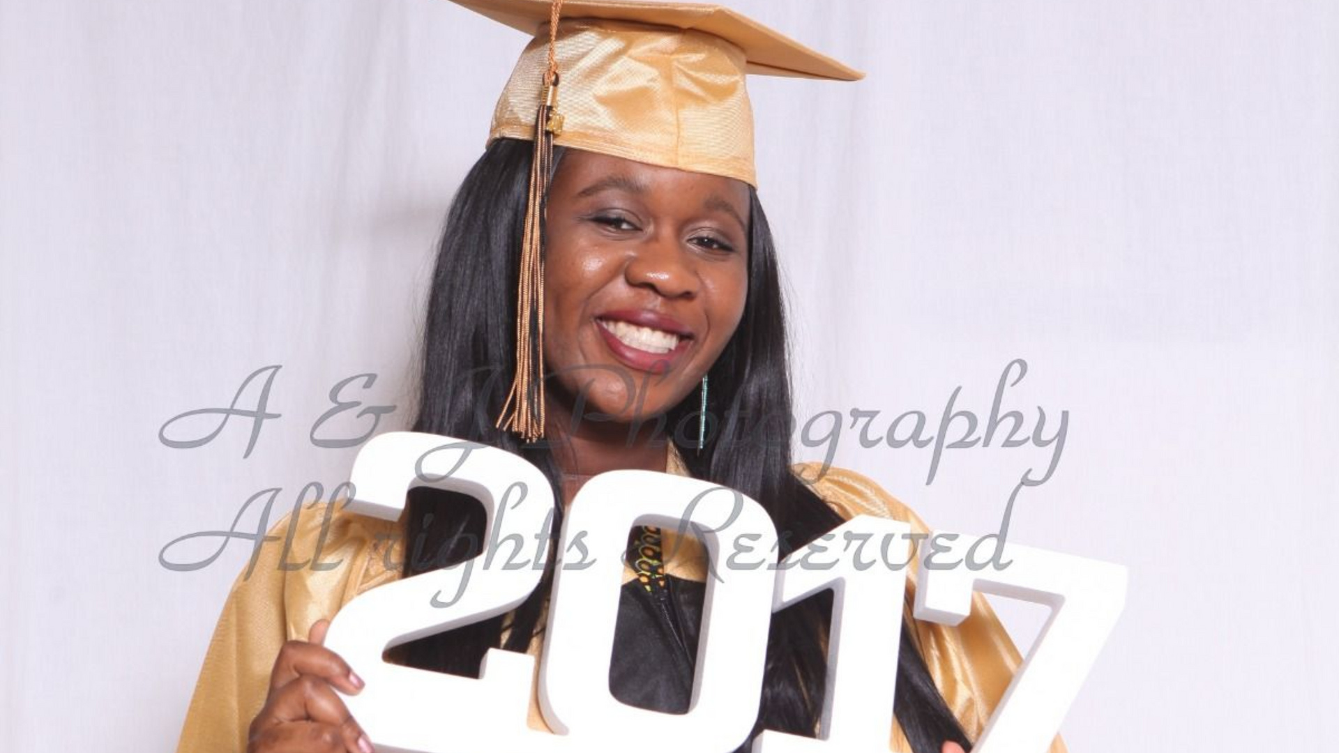 Woman in gold cap and gown holding a 2017 sign, smiling at a graduation backdrop.