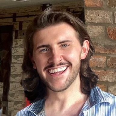 A man with long hair and a beard is smiling in front of a brick wall.
