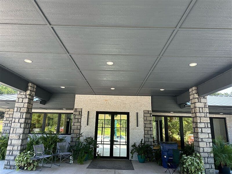 A low-angle view of a building entrance with a gray ceiling, stone columns, glass double doors, and potted plants.