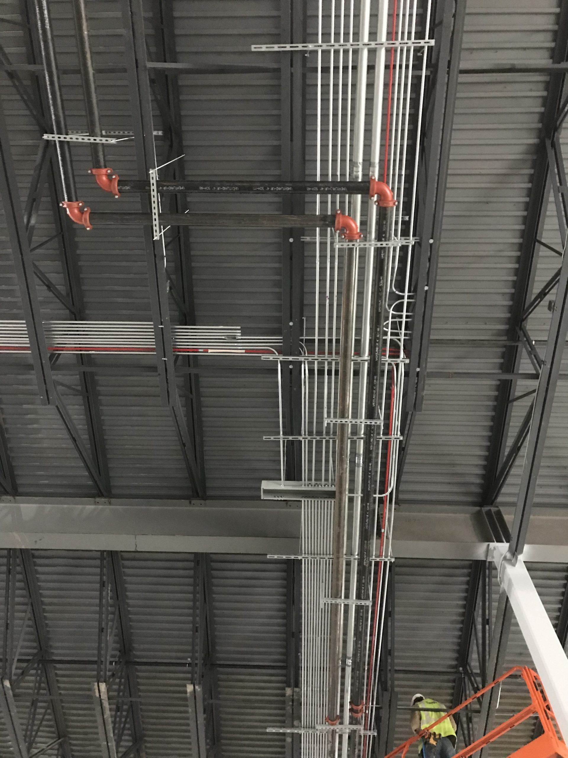 An industrial ceiling with numerous conduit pipes running along metal trusses, with a worker on a lift visible below.