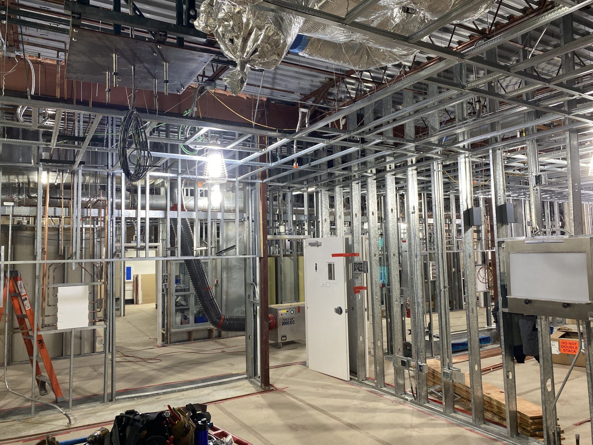 Interior view of a commercial building under construction with metal wall framing, exposed ceiling pipes, and a door.