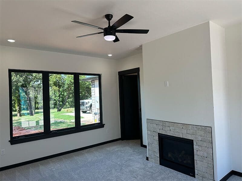 A living room with a large window, a ceiling fan, carpeted floor, and a stone-faced fireplace on a light-colored wall.