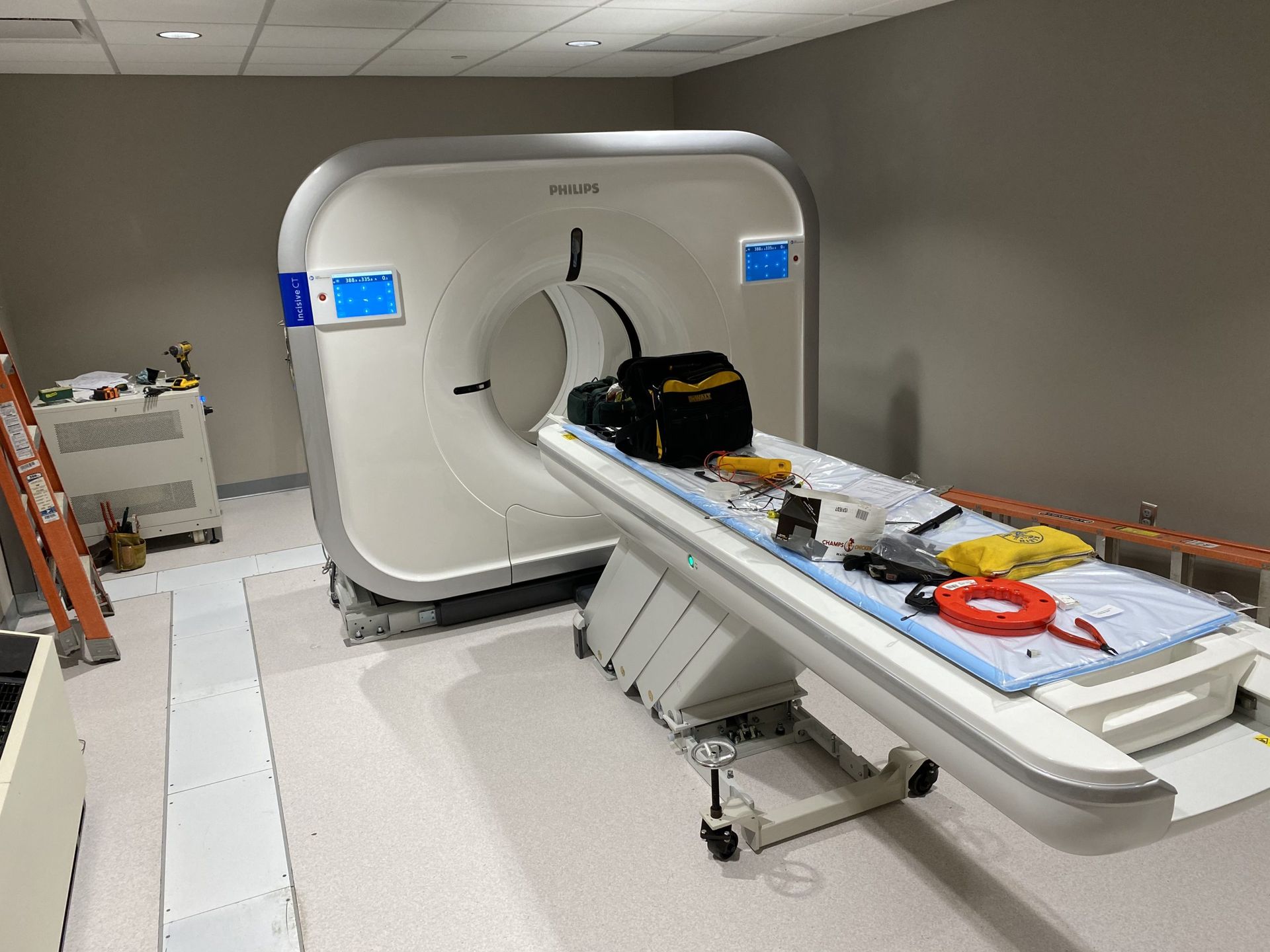 A medical CT scanner in a room with a patient bed holding a tool bag, medical equipment, and an orange ladder nearby.