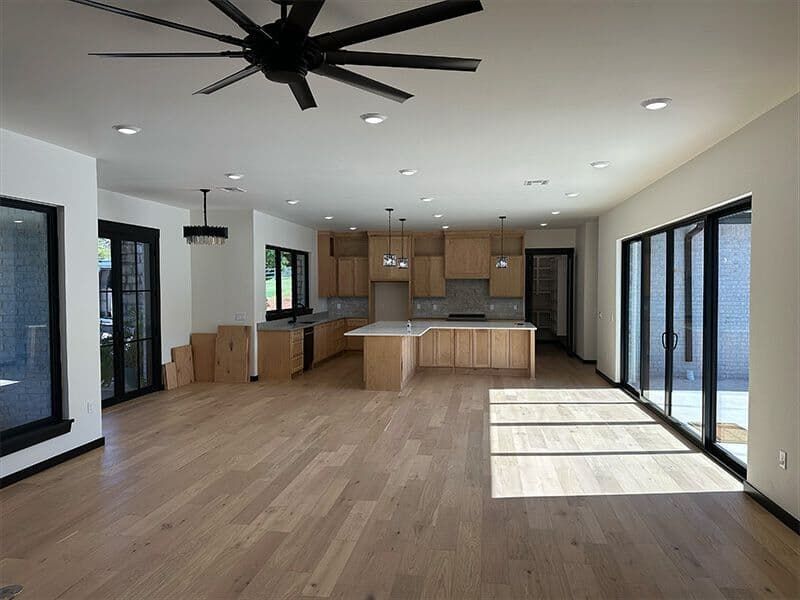 A spacious, modern kitchen and living area with light wood floors, natural wood cabinets, and large black-framed windows.