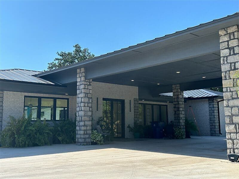 A modern single-story building with a large stone-pillared carport over a paved driveway under a clear blue sky.