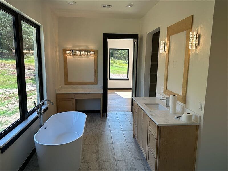 A modern bathroom with a freestanding white tub, wooden vanity, vanity desk, and a doorway leading to an outdoor view.