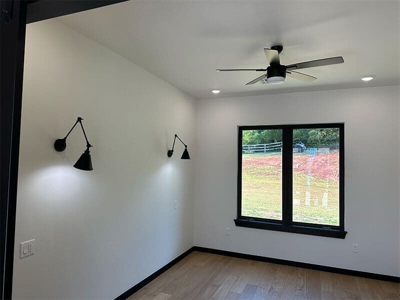 Minimalist room with white walls, wood floors, two black wall sconces, a black-framed window, and a ceiling fan.