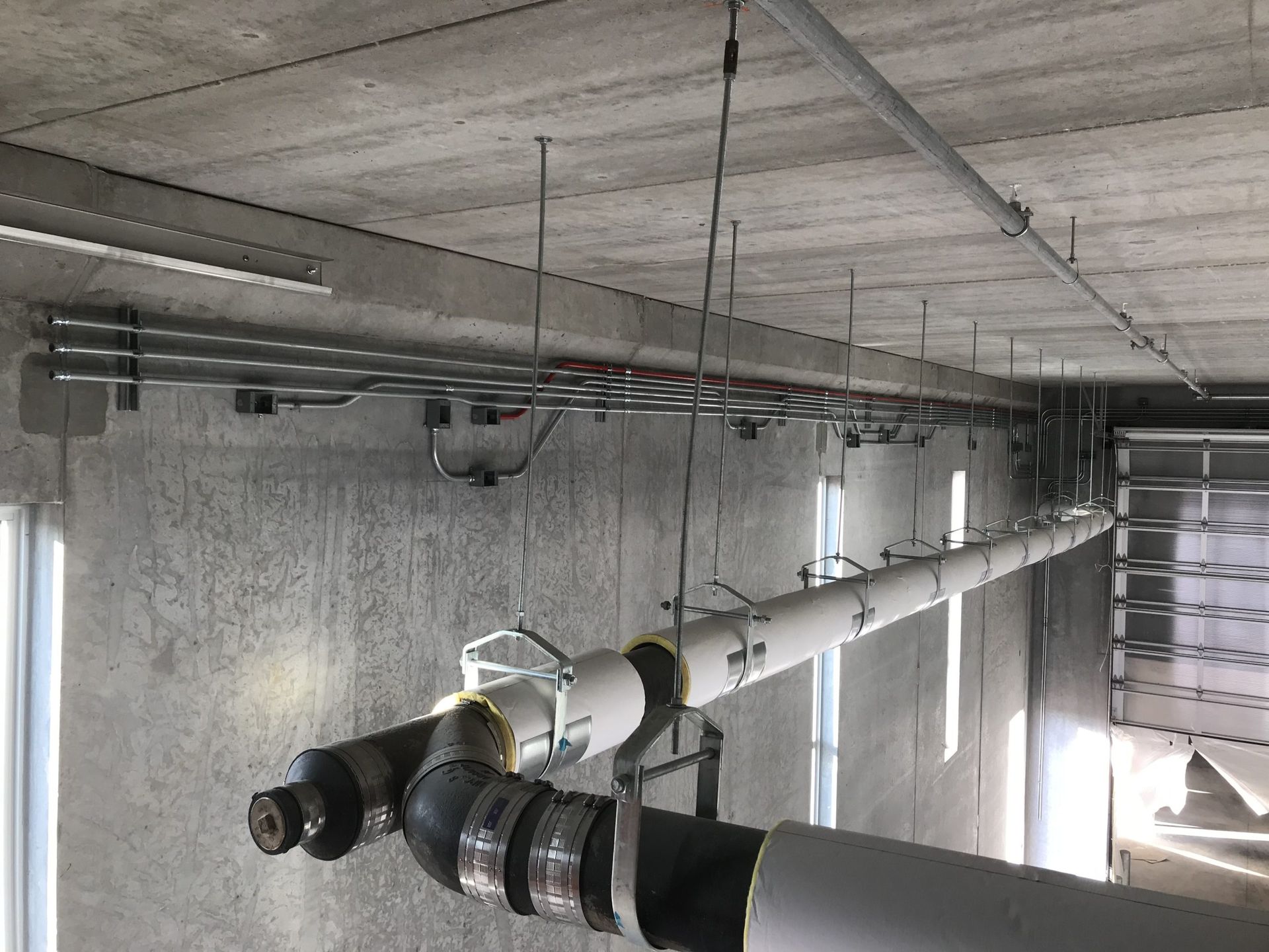 Insulated pipes and metal conduit runs suspended from a concrete ceiling in an industrial building interior.