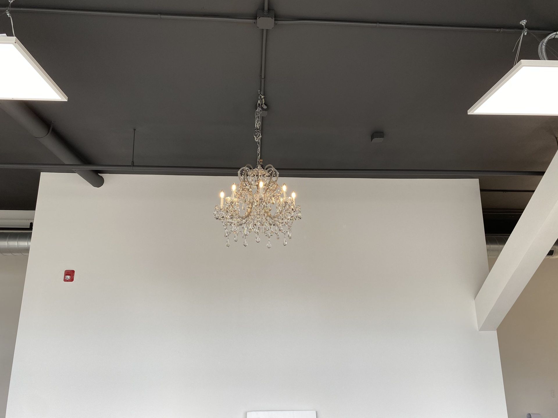 A crystal chandelier hangs in the center of a room with a dark ceiling and white wall, flanked by two square lights.