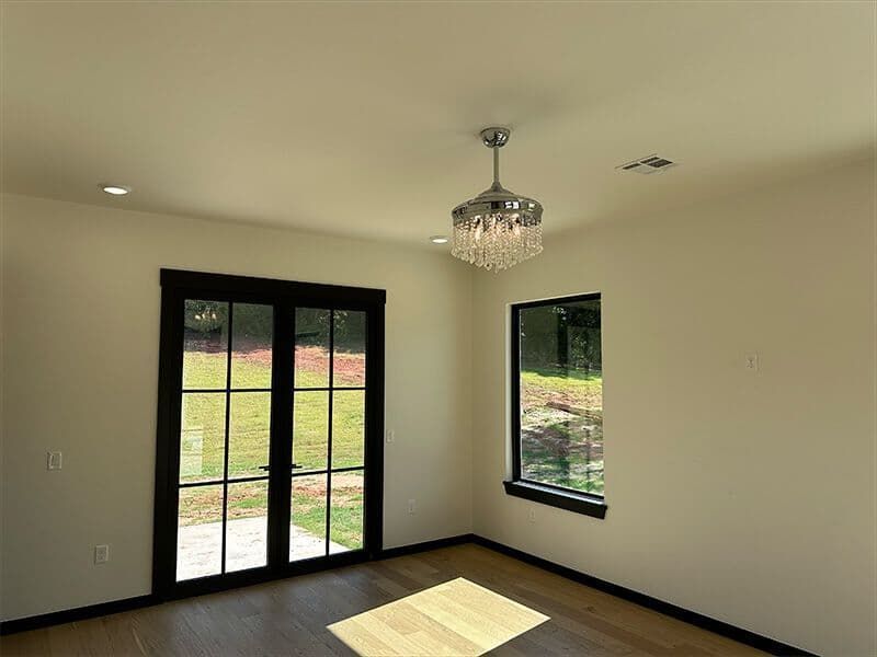 A bright, modern room with off-white walls, dark-framed French doors, a window, wood flooring, and a crystal ceiling light.