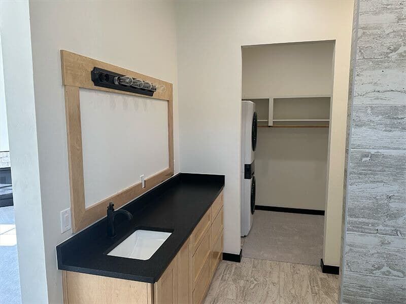 A laundry area with a light wood vanity, black countertop, sink, mirror, and a stacked washer/dryer in a walk-in closet.
