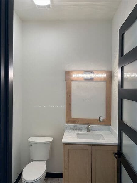 A small bathroom featuring a light wood vanity, white marble countertop, square mirror with wood frame, and white toilet.