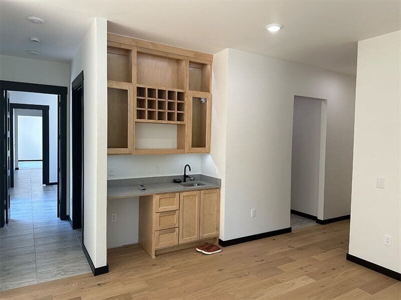 A wet bar with light wood cabinets, a wine rack, and gray countertops, situated in a hallway near a doorway.