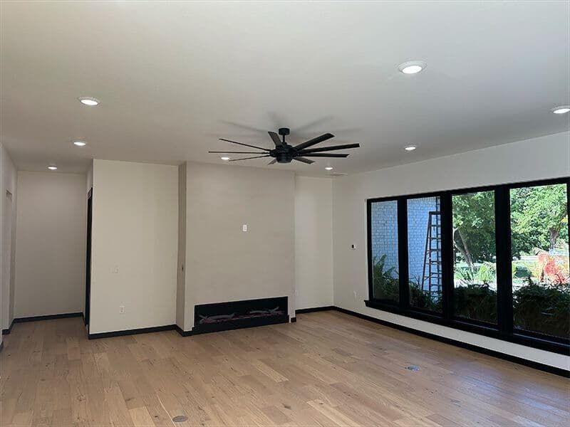 A bright, modern living room featuring light wood flooring, a fireplace, a black ceiling fan, and a large window wall.