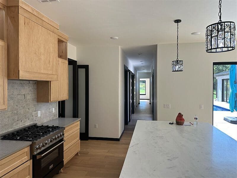 A bright modern kitchen with light wood cabinets, a marble island, a gas stove, and a long hallway leading to a patio.