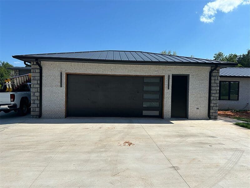 A modern garage with white stone walls, a dark metal roof, a wide door with side windows, and an adjacent entry door.