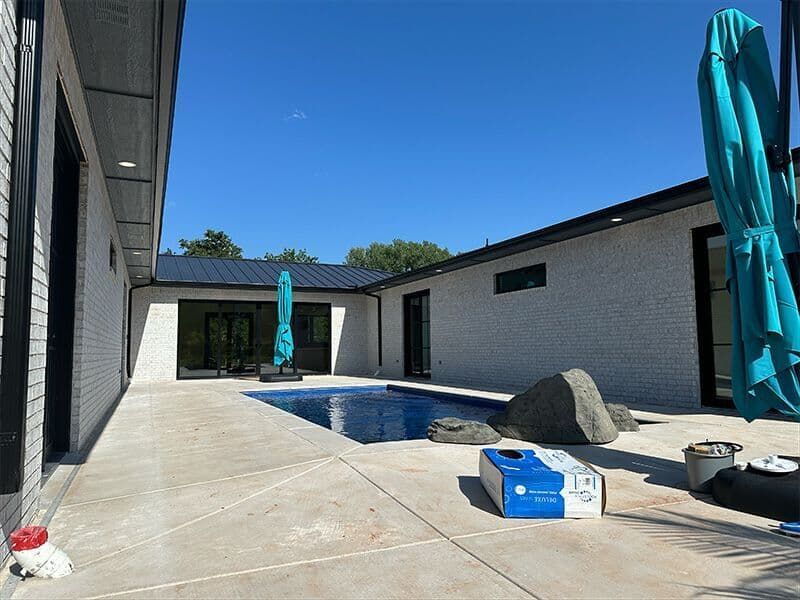 A white brick house surrounds a rectangular swimming pool with a concrete patio, large rocks, and a closed blue umbrella.