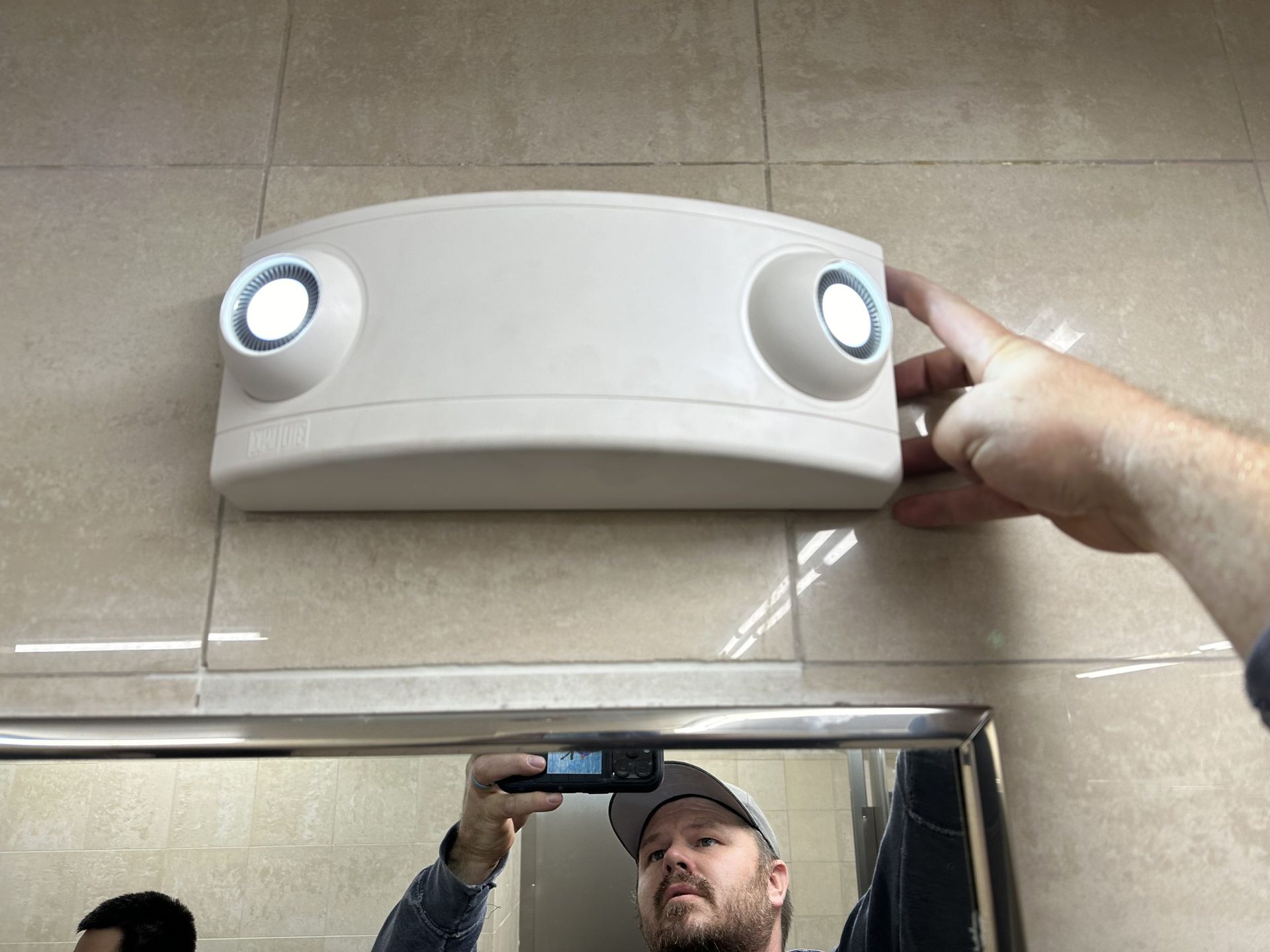 A person’s hand touching a wall-mounted emergency light above a mirror in a tiled room.