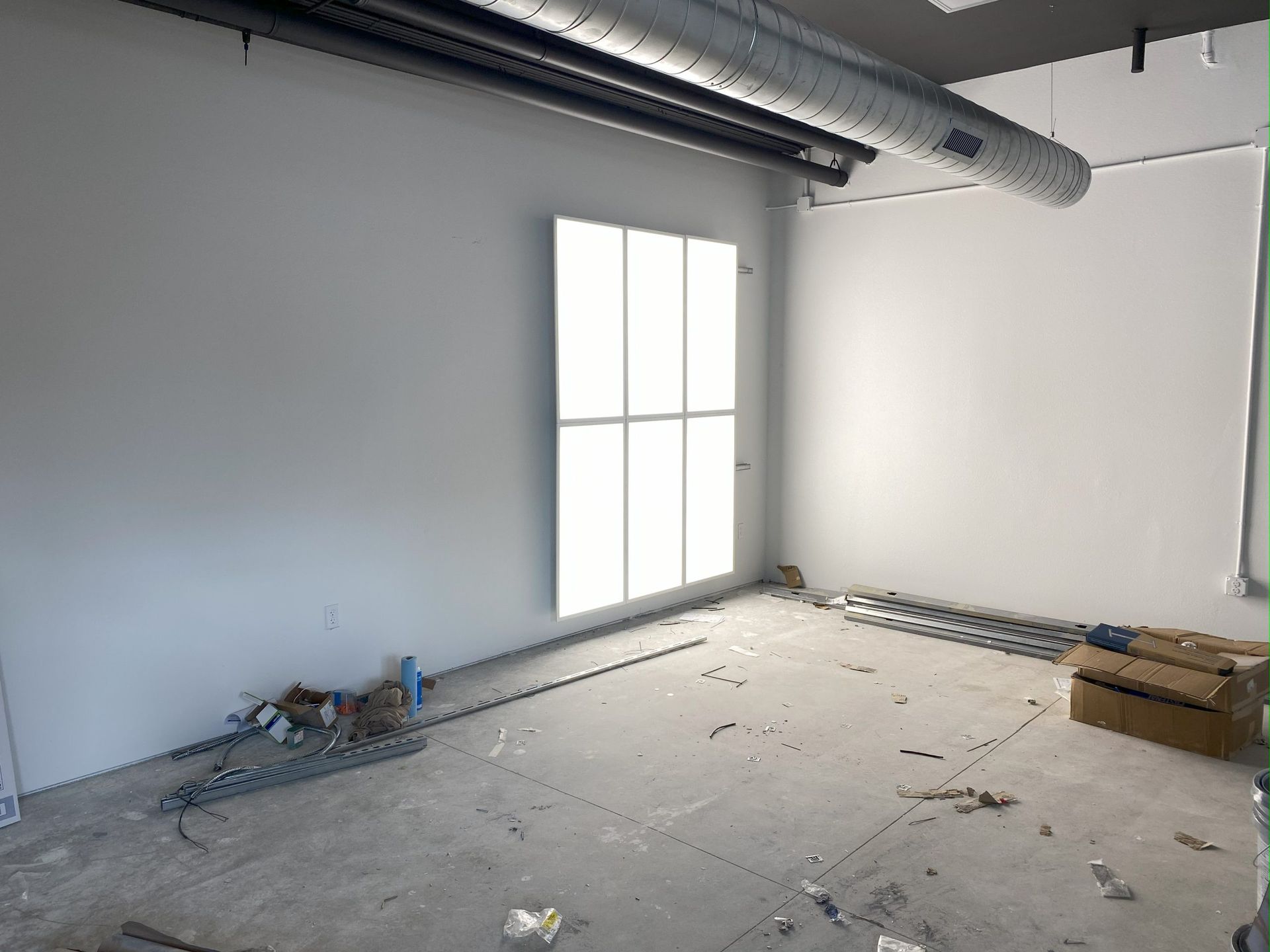 An empty, unfinished room with white walls, concrete flooring with debris, a bright light panel, and an exposed ceiling.