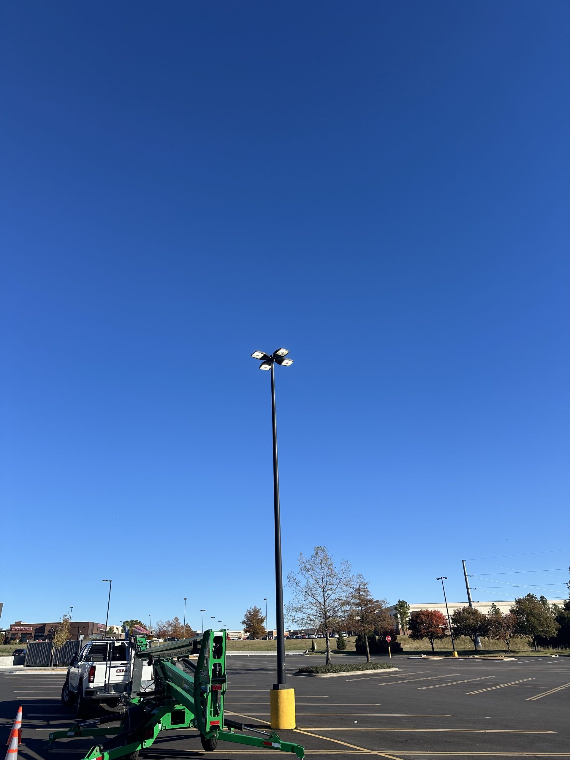 A tall parking lot light pole stands in an empty, paved lot under a clear blue sky, next to green construction equipment.