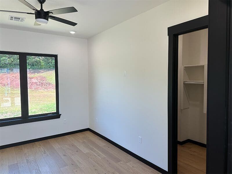 An empty bedroom with white walls, light wood floors, a ceiling fan, and a walk-in closet with black door trim.
