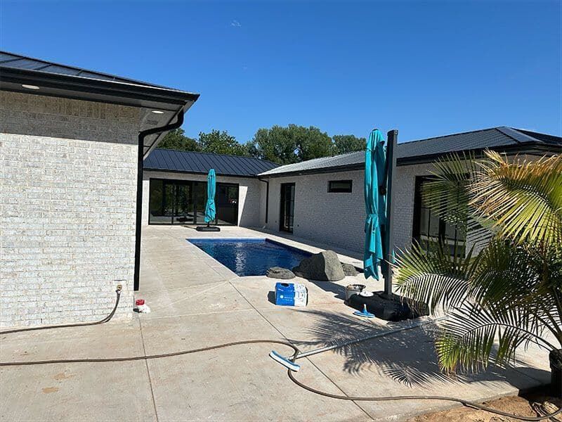A backyard features a rectangular swimming pool surrounded by a concrete patio, white brick exterior walls, and umbrellas.