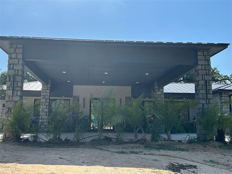 A modern house entrance with a large covered porch, stone columns, dark grey ceiling, and palm trees against a blue sky.