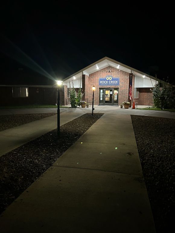 A brick building with a brightly lit entrance at night, approached by a concrete walkway flanked by small lamps.