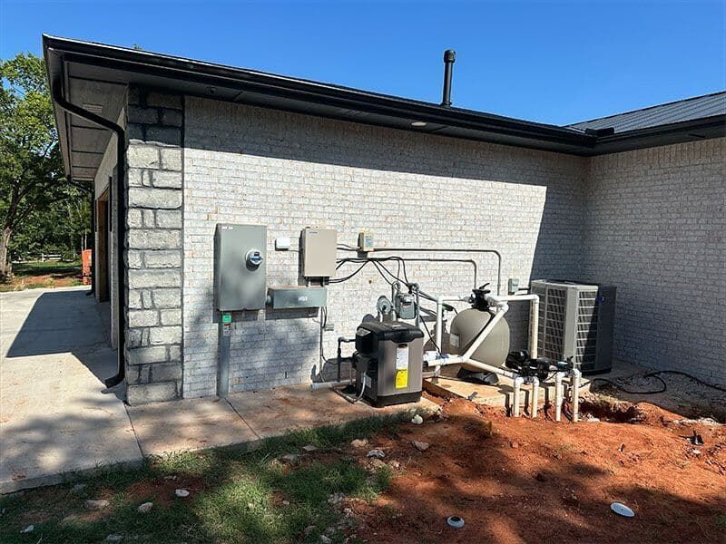 The exterior wall of a brick house featuring an electrical panel, a pool filter pump, and HVAC equipment on dirt ground.