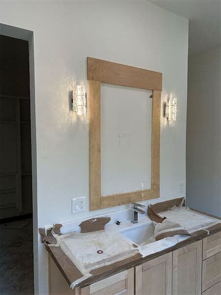 A light-colored wooden bathroom vanity with a mirror framed in matching wood, flanked by two wall-mounted light fixtures.