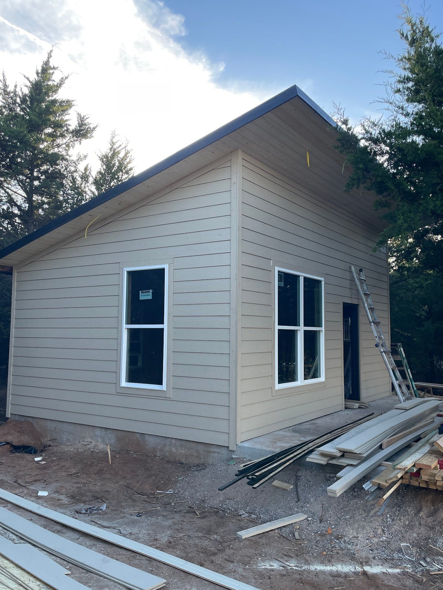 A tan, single-story shed under construction with two windows, a slanted roof, and materials scattered on the ground.