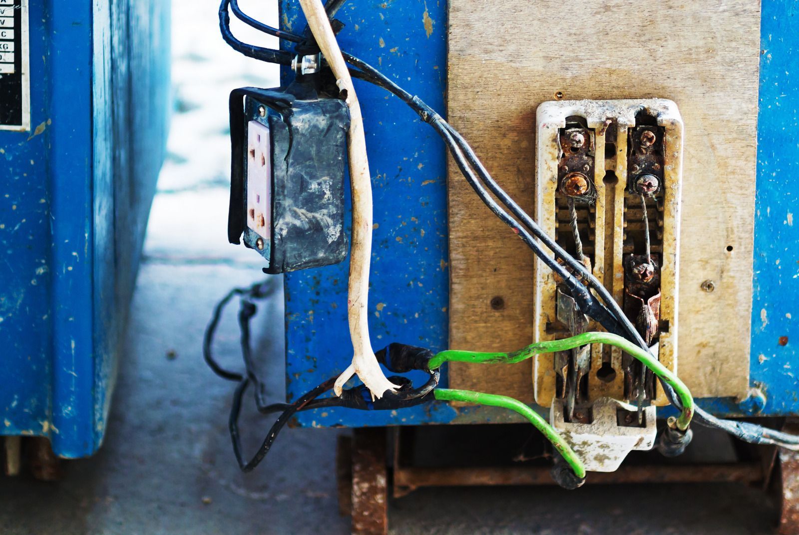 Unsafe, exposed electrical wiring and connections mounted on a wooden board attached to a blue metal frame.
