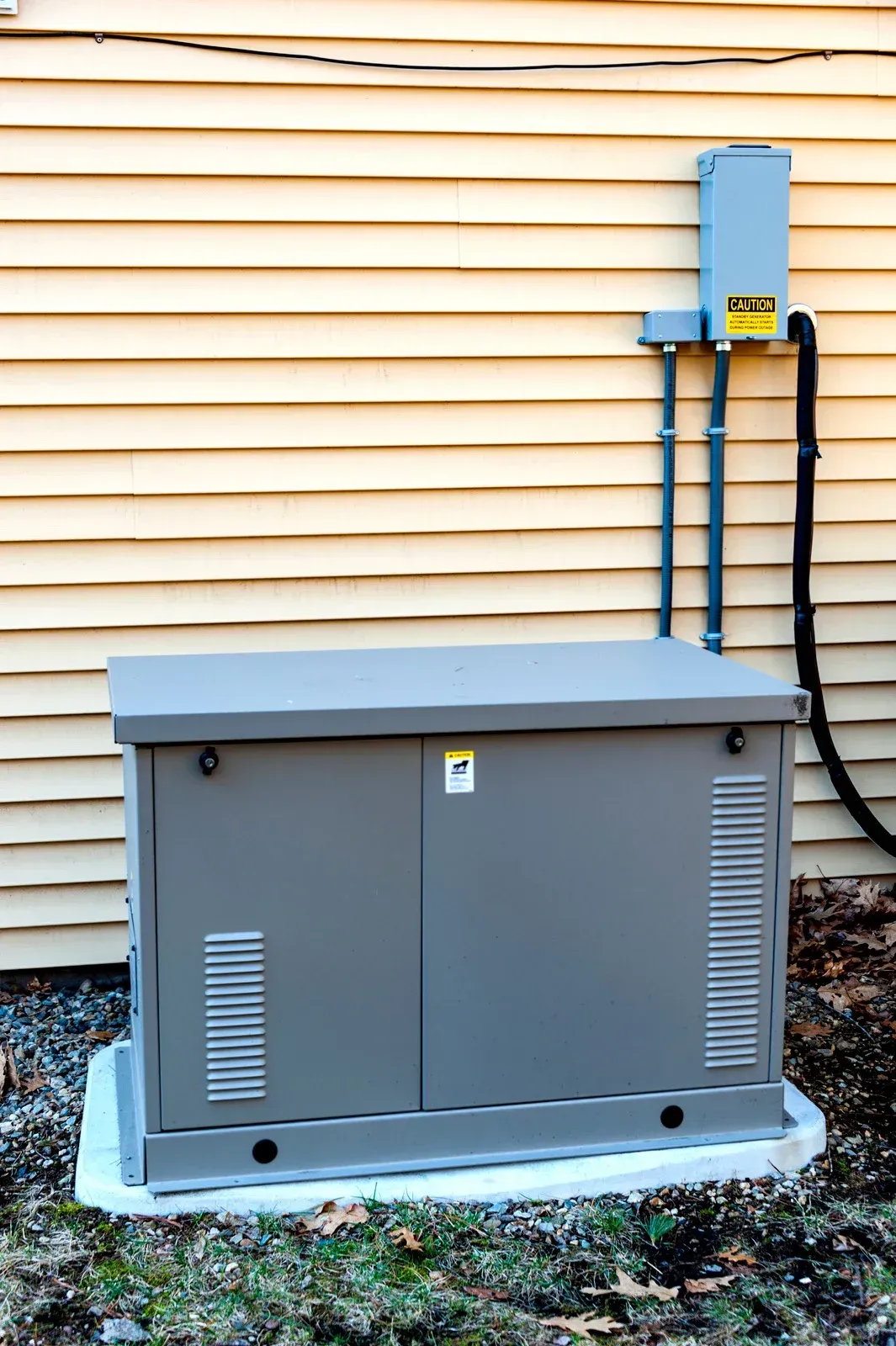 A grey standby home generator sits on a concrete pad against a yellow-sided house with external electrical equipment.