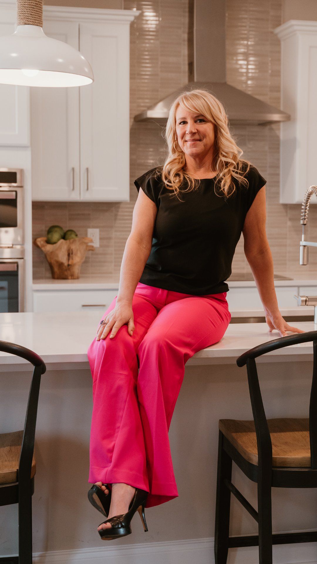 Woman in black top and bright pink pants sitting on a kitchen island, smiling.