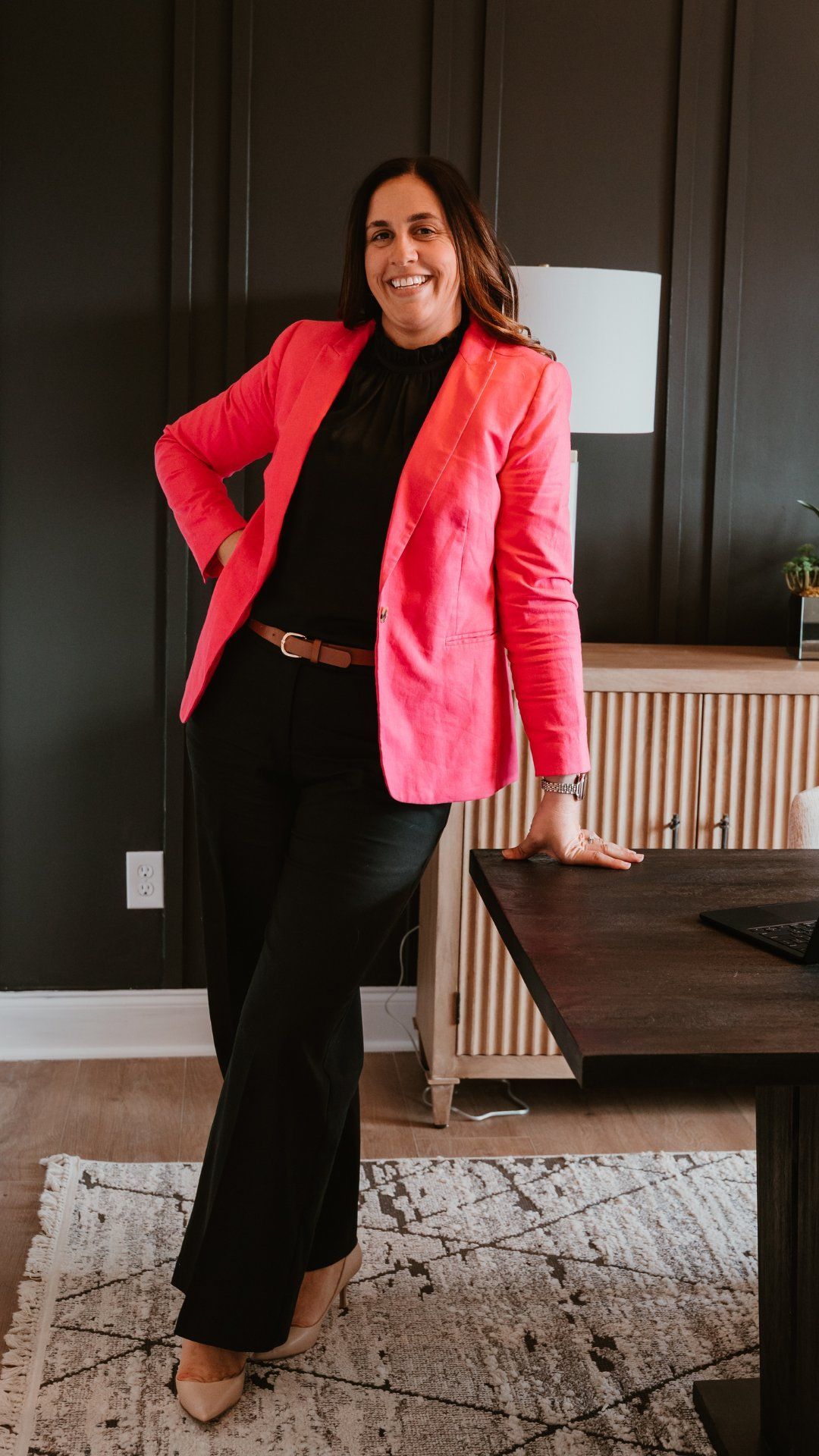 Person in a bright pink blazer and black outfit leaning on a table in a dark-paneled room.