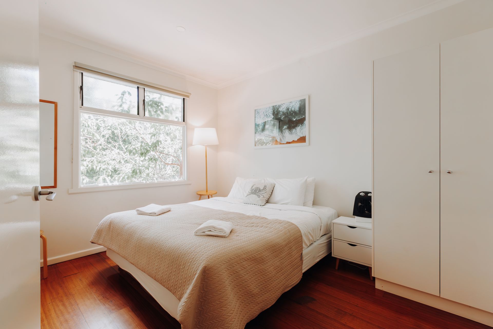 A bright bedroom with a bed featuring a textured beige cover, a white wardrobe, a bedside table, and a window with light.