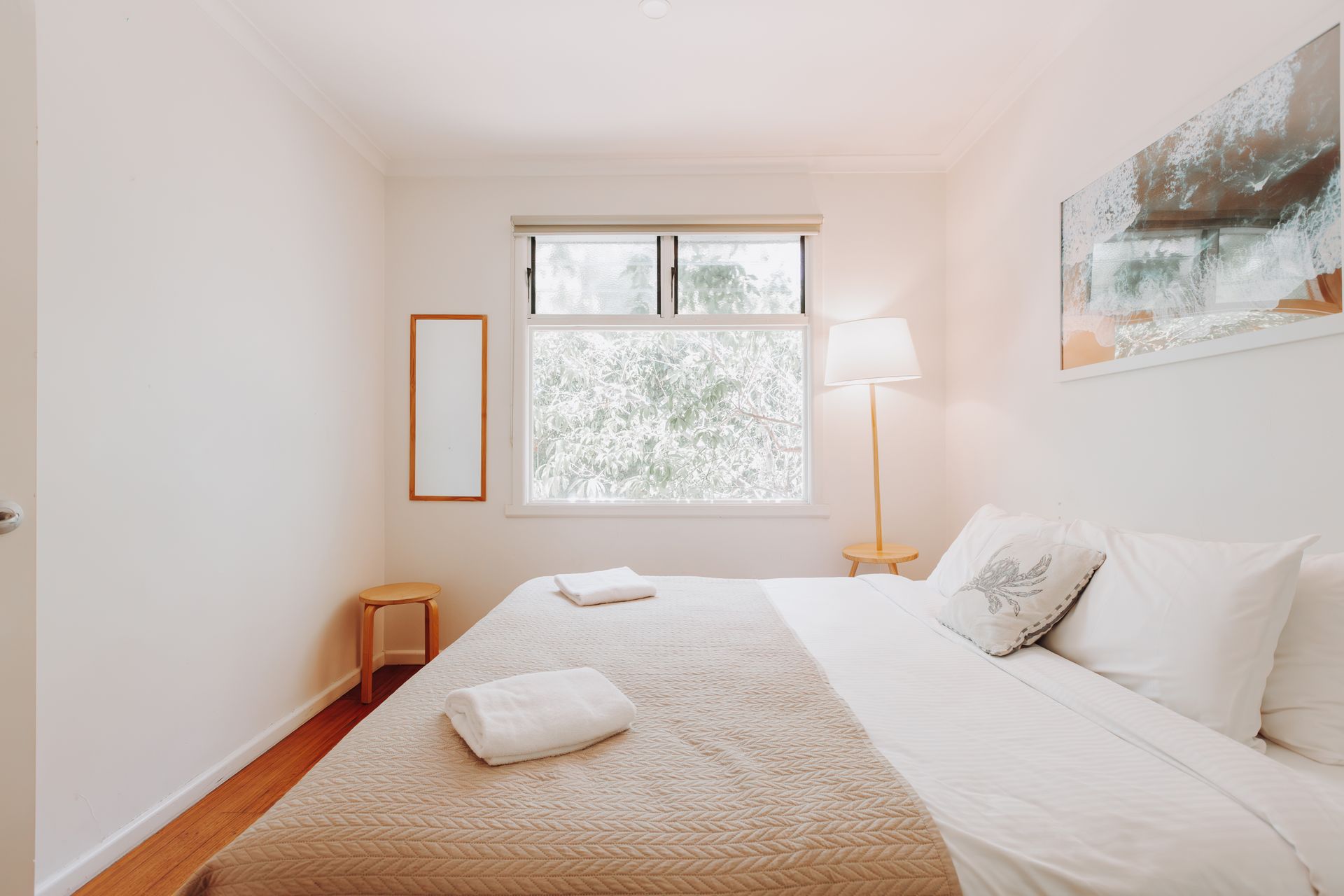 A bright bedroom featuring a bed with a tan textured blanket and white pillows, a floor lamp, and a window with trees.