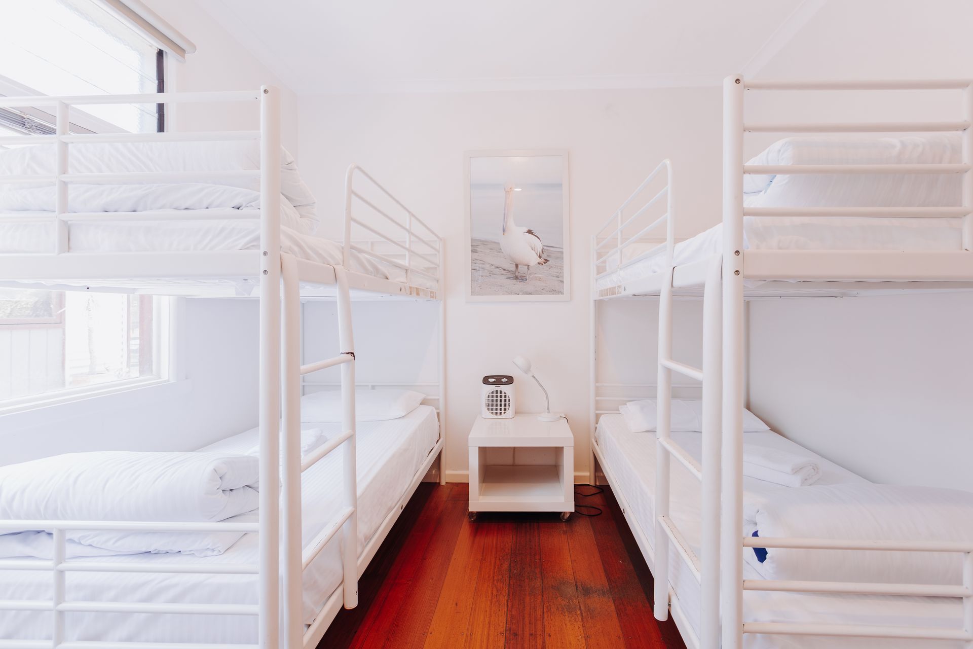 A minimalist hostel room with two white metal bunk beds on opposite sides, wooden floors, and a small nightstand.