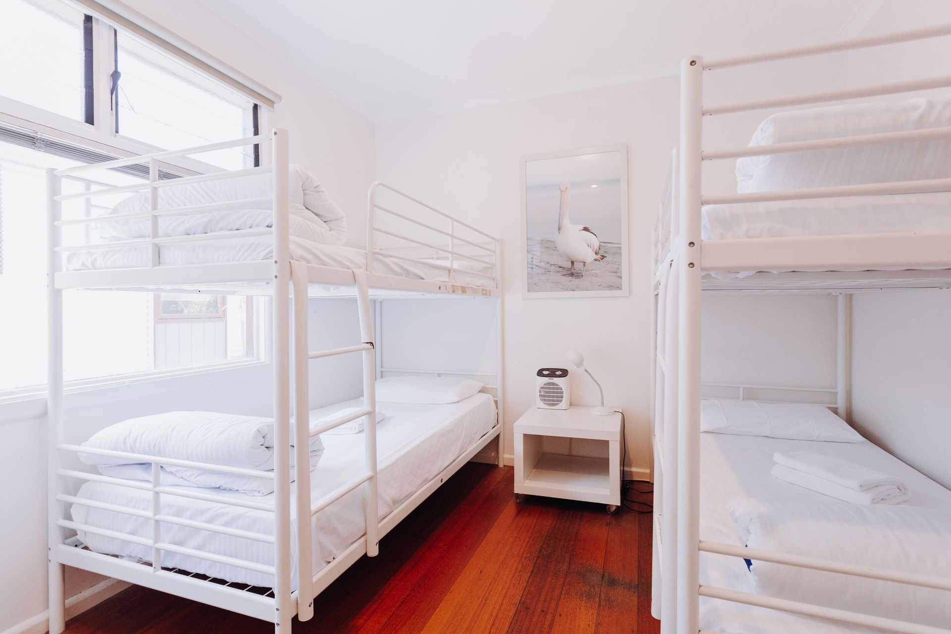 Two white metal bunk beds in a room with white walls, a nightstand, and dark wooden floors.