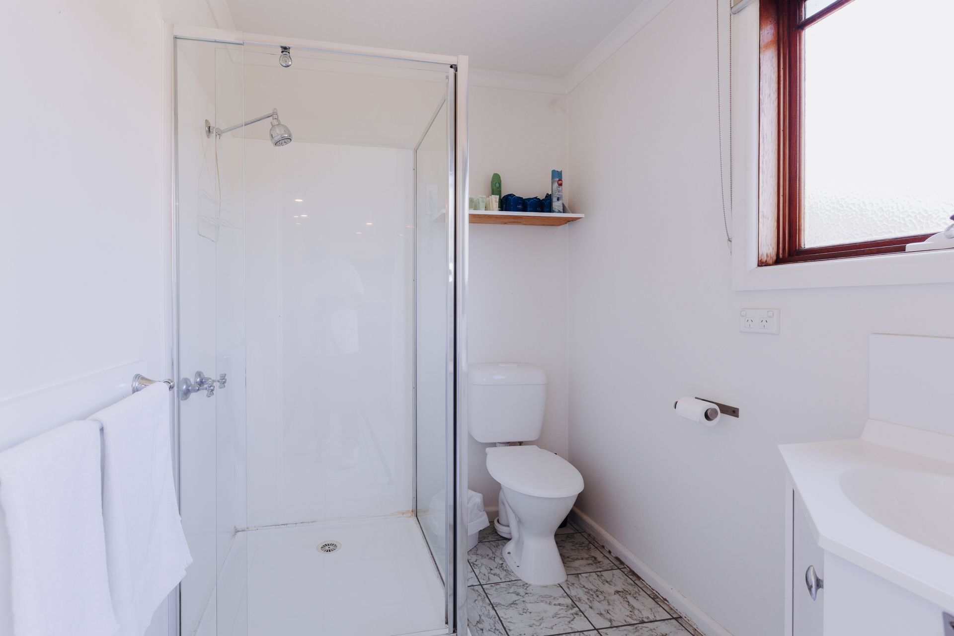 A bright, minimalist bathroom with a white shower enclosure, a toilet, a small wooden shelf, and patterned floor tiles.