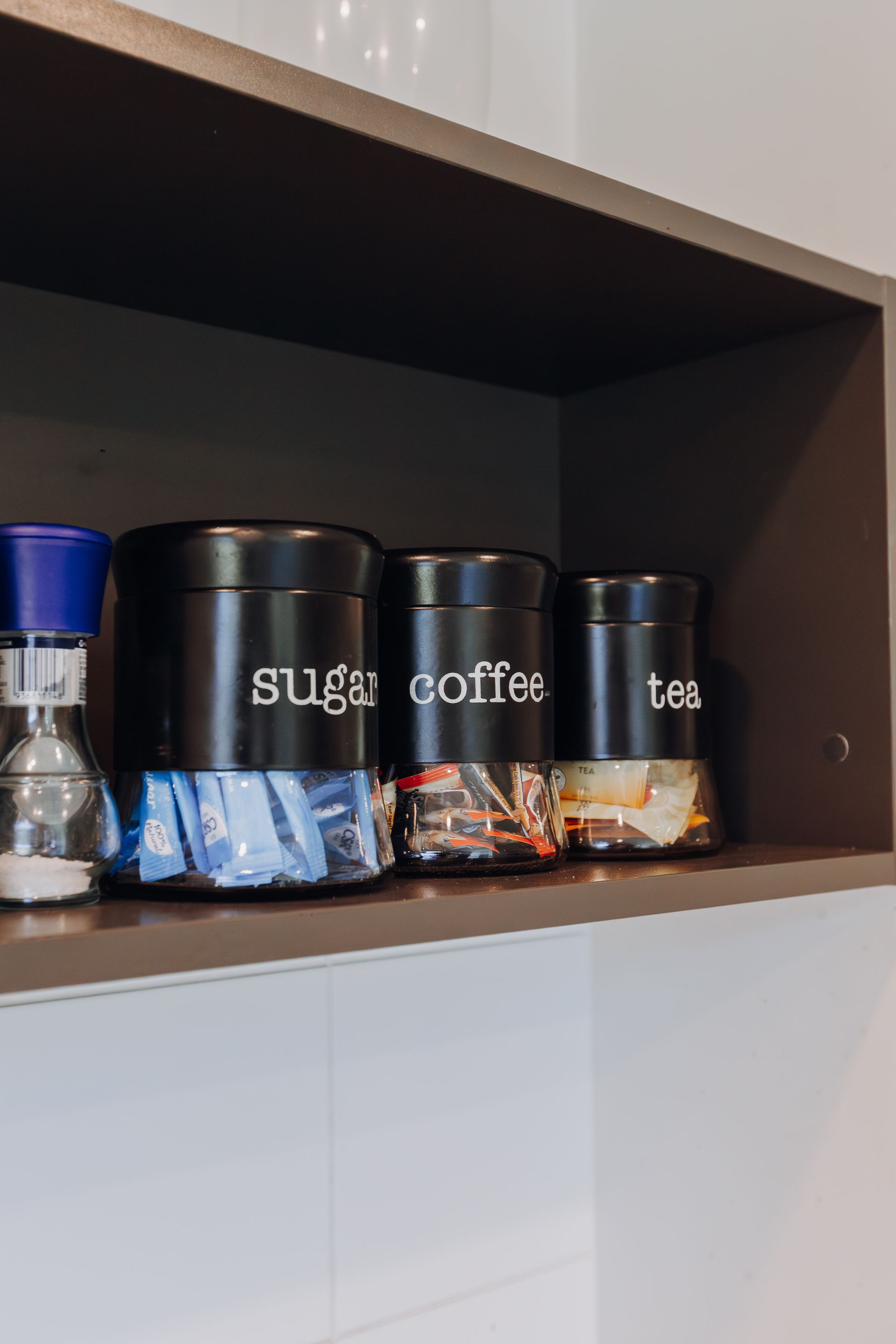 Three black labeled canisters for sugar, coffee, and tea sit on a shelf next to a salt or pepper shaker.