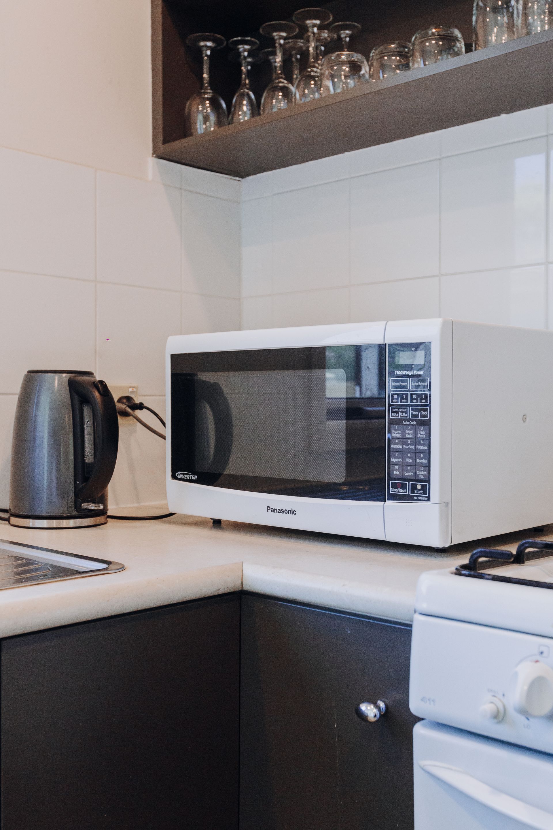 A kitchen counter with a gray electric kettle, white microwave, and shelves with wine glasses above.