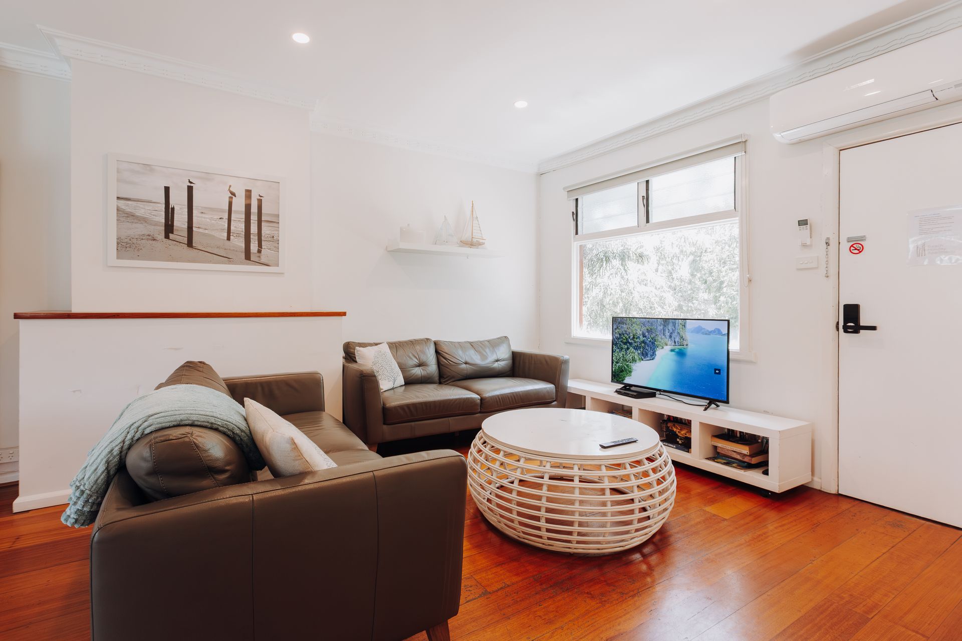 A cozy, brightly lit living room with wooden floors, two dark grey sofas, a white wicker coffee table, and a TV.