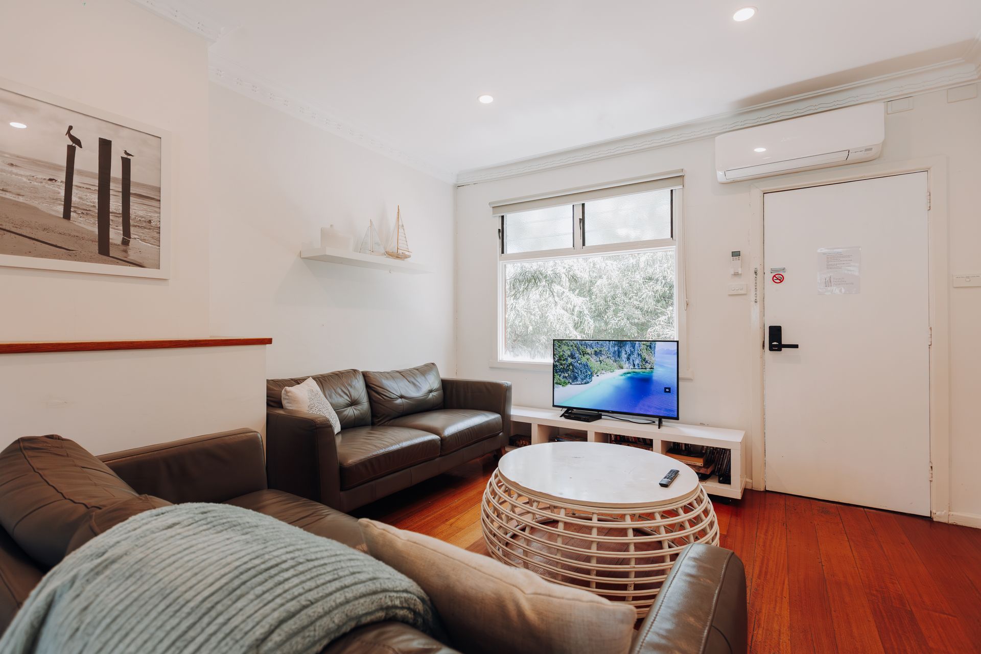 A cozy living room with leather sofas, a wicker coffee table, a TV on a stand, and a white door with an air conditioner.