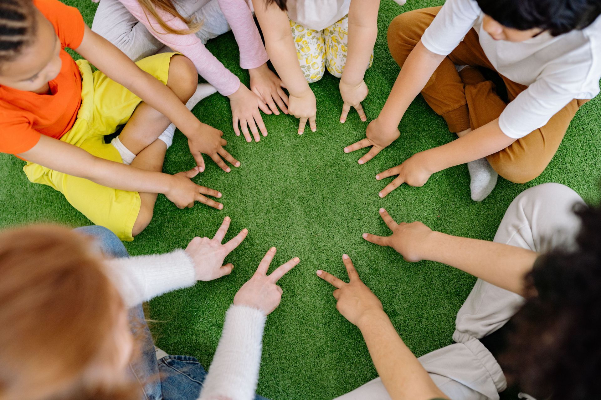 Children sitting in a circle on grass, reaching hands toward the center.