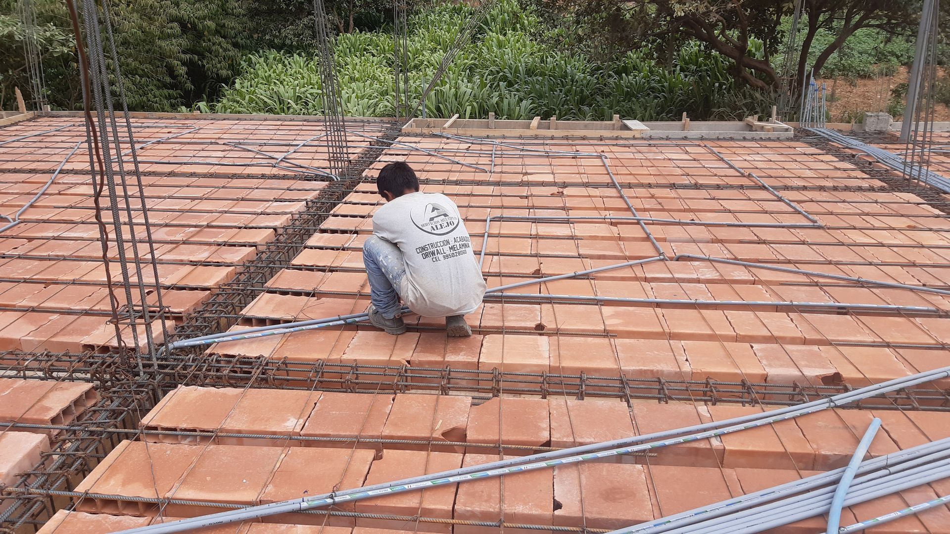 Un trabajador se encuentra agachado sobre una rejilla de varillas de refuerzo en un tejado en construcción, con árboles al fondo.
