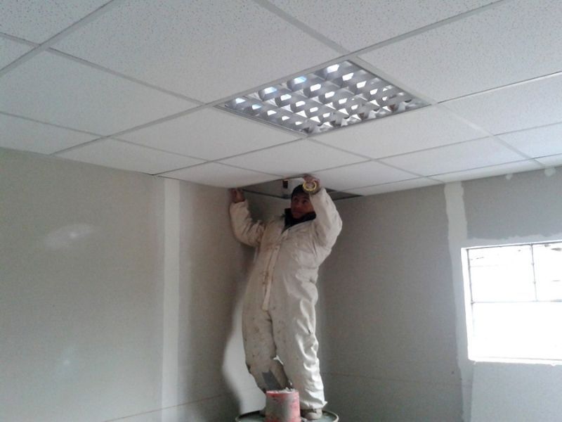 Trabajador con mono blanco instalando o reparando un panel de techo en una oficina.