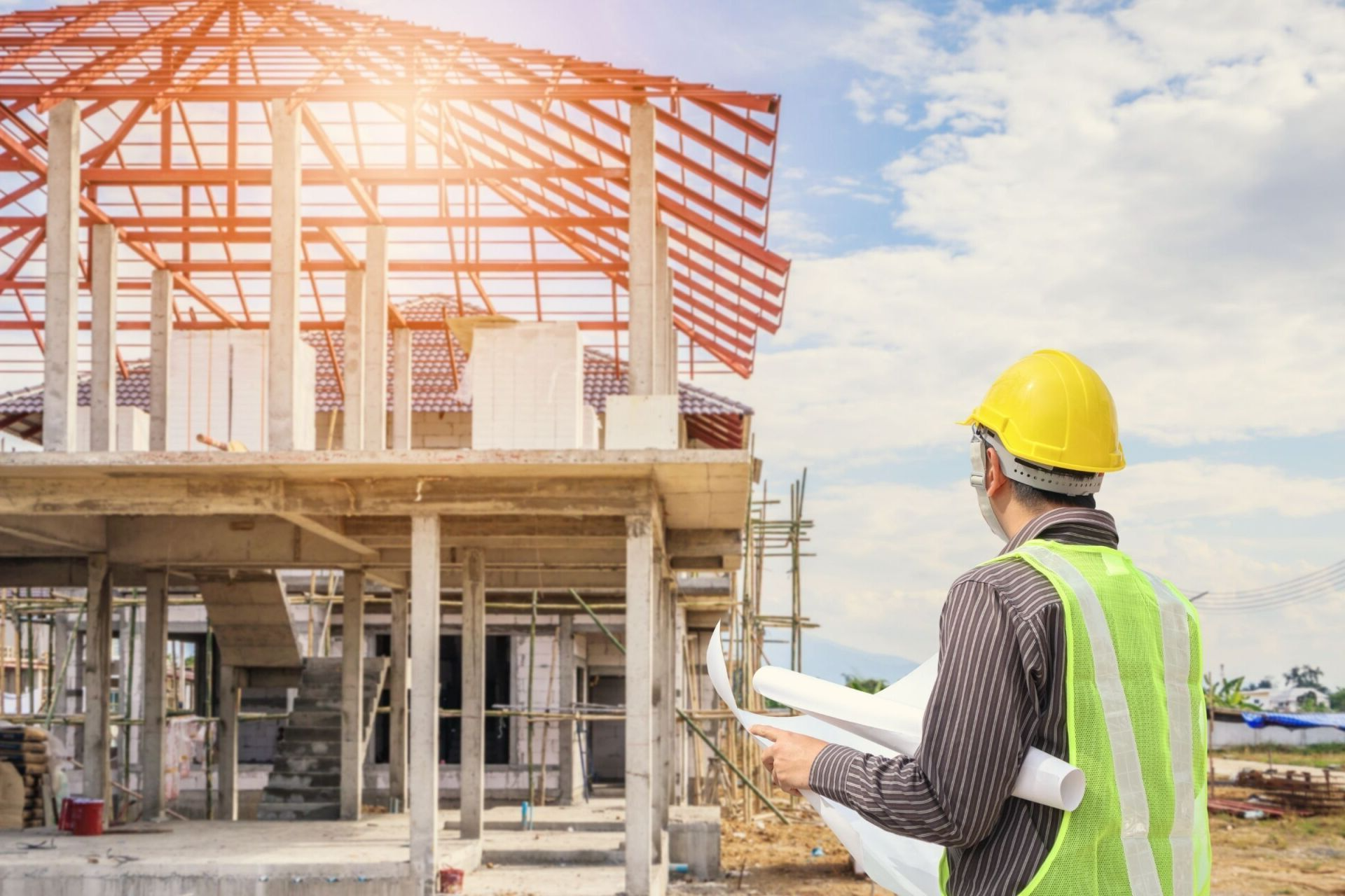 Obra en construcción con un trabajador con casco amarillo inspeccionando la estructura de un edificio con planos en la mano.